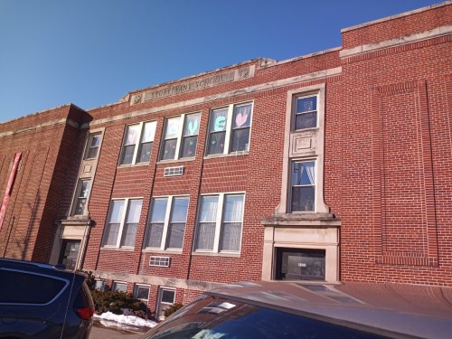 Brick school building with colorful "LIVE" signs in the windows, under a clear blue sky. Snow on the ground.