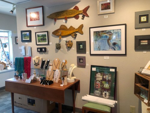 A gallery wall displays various artworks, including fish sculptures, framed prints, and handmade jewelry on a table.