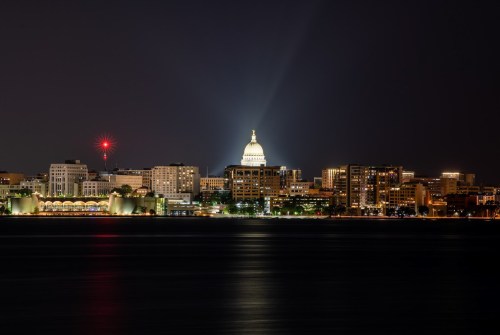 Night view of a city skyline featuring a prominent dome building, illuminated against a dark sky with reflections on water.