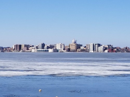 City skyline of Madison, Wisconsin, with frozen lake in the foreground and clear blue sky above.