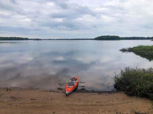 A bright orange kayak rests on a sandy shore by a calm lake, surrounded by greenery and cloudy skies.