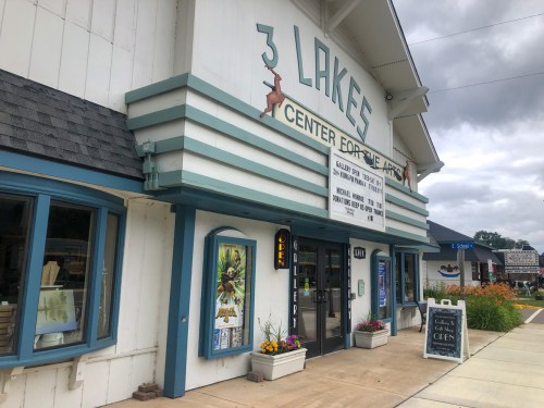 Exterior of the 3 Lakes Center for the Arts, featuring a marquee and colorful flower beds, under a cloudy sky.