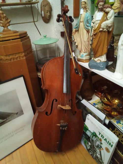 A vintage cello stands among various antiques and figurines in a cluttered display.