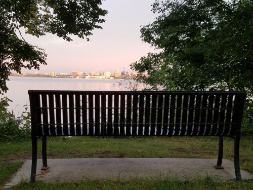 A black bench facing a calm lake, with a city skyline visible in the background under a soft sunset glow.