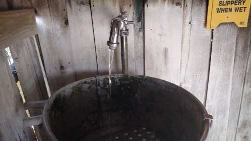 A metal faucet pours water into a large, weathered metal basin against a wooden wall. A caution sign is visible nearby.