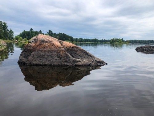 A large rock partially submerged in calm water, reflecting the cloudy sky and surrounding trees.