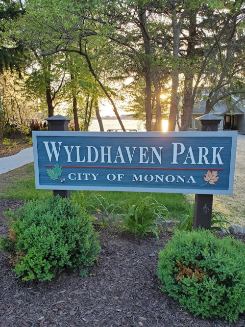 Sign for Wyldhaven Park in Monona, surrounded by greenery, with a sunset view over the water in the background.