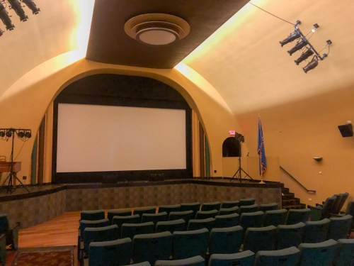 Interior of a vintage theater with a large screen, green seats, and a curved ceiling. Stage lights are visible.