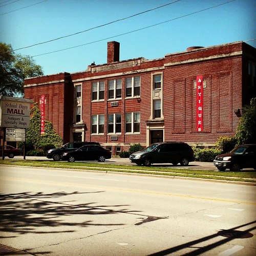 A brick building with "Antiques" signs, located across from a street with parked cars and trees in the background.