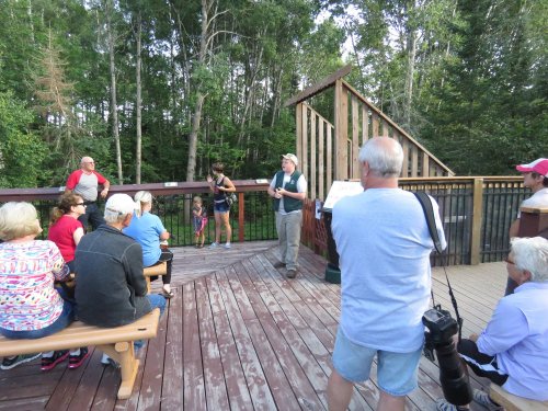 A group of people gathers on a wooden deck in a forested area, listening to a speaker.