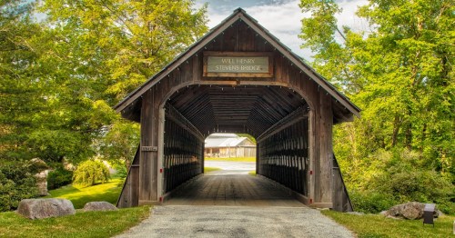 A wooden covered bridge with a sloped roof, surrounded by greenery and rocks under a cloudy sky.