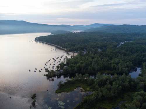 Aerial view of a serene lake surrounded by lush forests and boats moored along the shore under a cloudy sky.