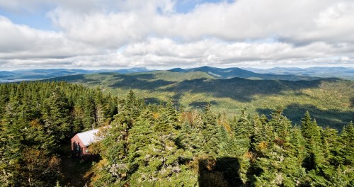A scenic view of mountains and forests under a cloudy sky, with a small cabin nestled among the trees.