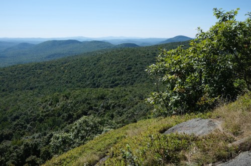 A panoramic view of lush green mountains under a clear blue sky, with a rocky foreground and distant hills.