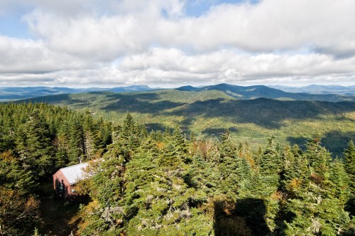 A scenic view of rolling green mountains and a small cabin, under a partly cloudy sky.