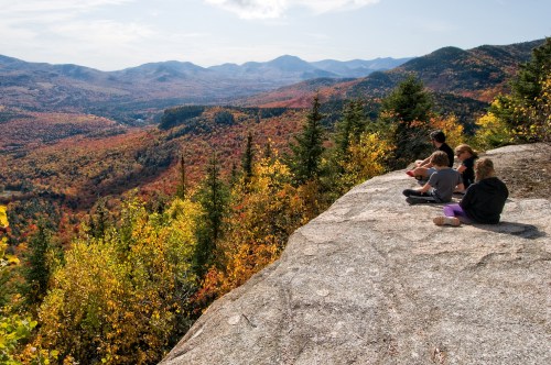 Four people sit on a rocky ledge, overlooking a vibrant autumn landscape of mountains and colorful foliage.