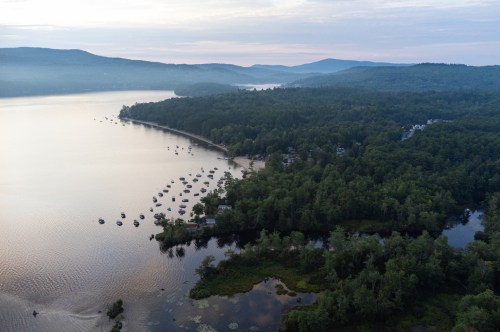 Aerial view of a serene lake surrounded by lush forests and mountains, dotted with boats along the shoreline.