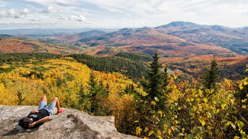 A person relaxes on a rock, overlooking a vibrant autumn landscape of mountains and forests.