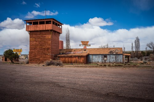 Abandoned building with a tall watchtower, surrounded by sparse vegetation and a cloudy blue sky.