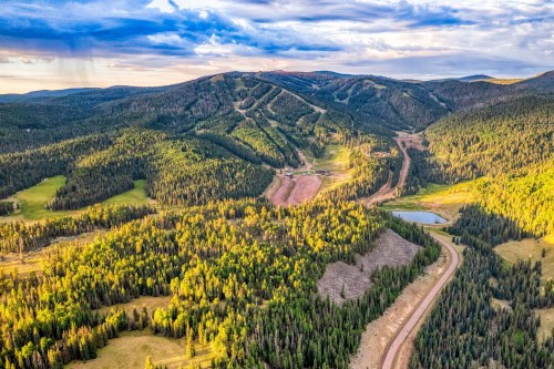 Aerial view of lush green mountains with a winding road, a small lake, and ski slopes in the distance under a cloudy sky.