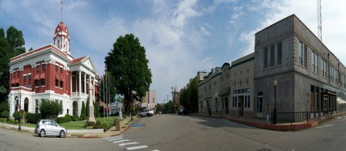 Panoramic view of a town square featuring a red brick courthouse and a historic building on a sunny day.