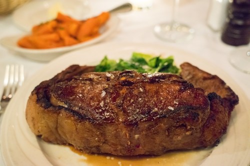 A juicy steak on a plate, garnished with salt, accompanied by greens and a side of carrots.