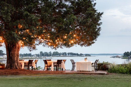 A serene outdoor dining area under a tree with string lights, overlooking a calm lake at dusk.