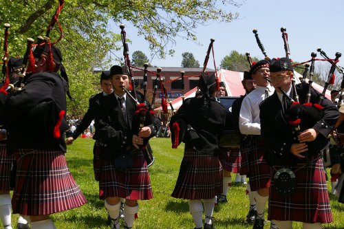 A group of bagpipers in traditional attire perform outdoors, surrounded by greenery and a festive atmosphere.