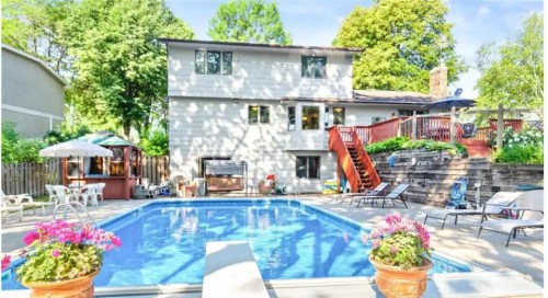 A backyard with a swimming pool, deck, and flowers, featuring a two-story house surrounded by trees.