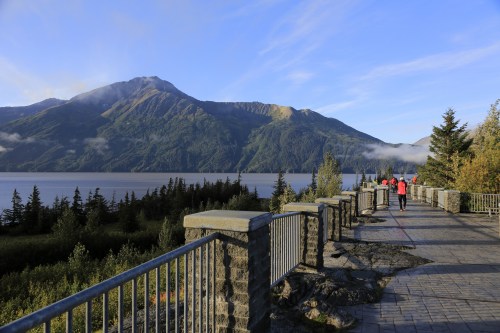 A scenic view of mountains and a lake, with people walking along a stone pathway surrounded by trees.