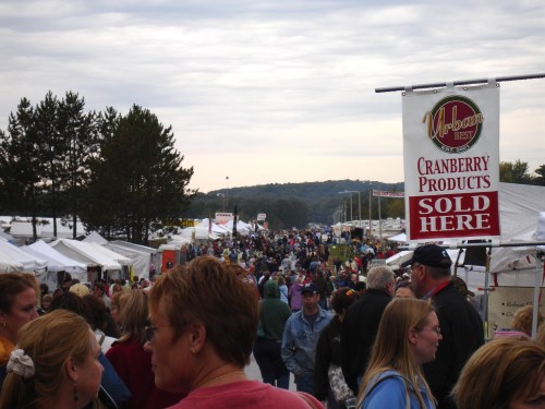 A bustling outdoor market with crowds of people, tents, and a sign advertising cranberry products.