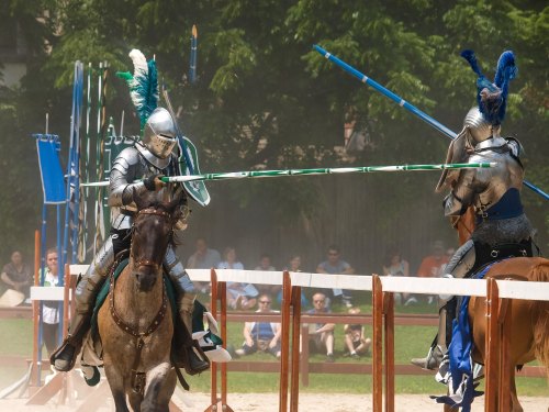 Two armored knights on horseback engage in a jousting match, with spectators watching in the background.