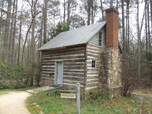 A rustic log cabin with a stone chimney, surrounded by trees in a wooded area. A sign reads "Paul Green Cabin."