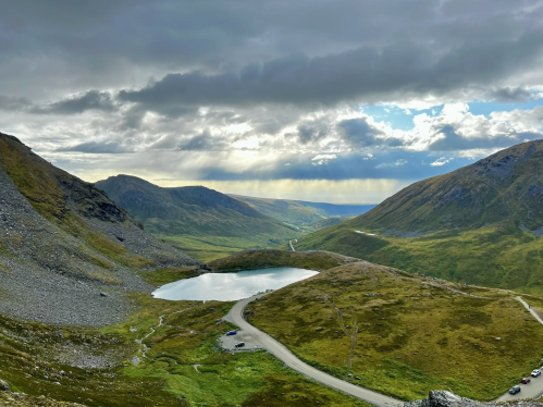 A scenic view of mountains and a tranquil lake under a cloudy sky, with a winding road in the foreground.