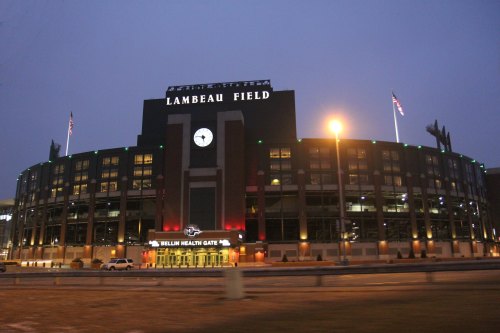 Lambeau Field at dusk, featuring the iconic clock and entrance, with flags and lights illuminating the exterior.