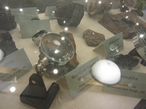 Display of mineral specimens under glass, featuring labels for goethite, hematite, and quartz on goethite.