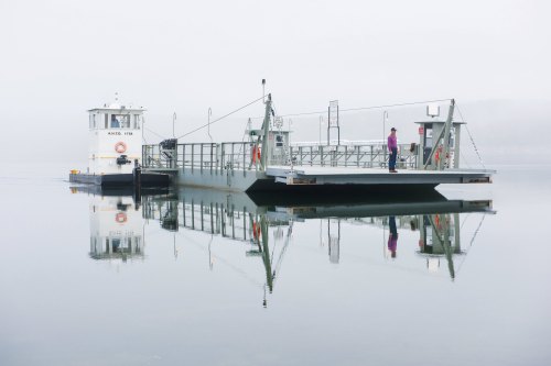 A ferry docked on calm water, with a person standing on the deck and misty surroundings reflecting in the water.