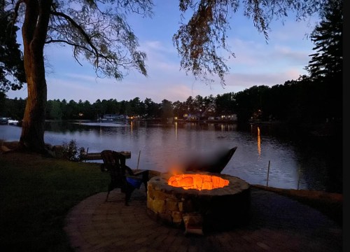 A serene lakeside scene at dusk, featuring a glowing fire pit and a calm water view surrounded by trees.