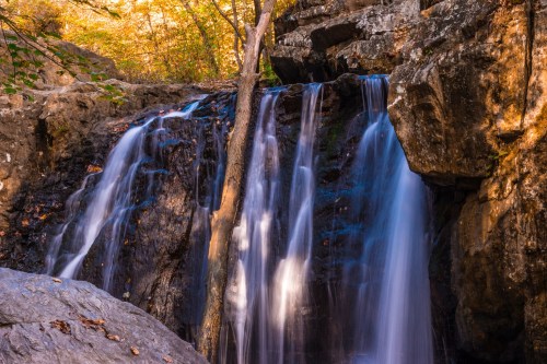 A serene waterfall cascading over rocks, surrounded by autumn foliage and trees in a tranquil forest setting.