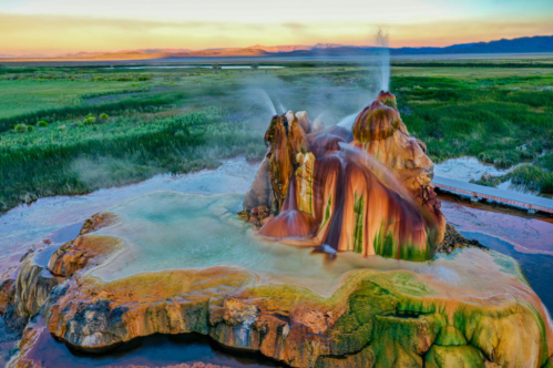 Aerial shot of colorful red, orange, yellow, and green Fly Geyser in Nevada in the Black Rock Desert. Water spews from the top. Green grasses and reeds surround it.