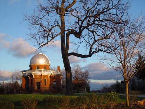 A historic observatory with a silver dome, surrounded by bare trees and a grassy area under a cloudy sky.