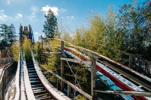 Abandoned roller coaster with overgrown greenery and snow, set against a clear blue sky and distant trees.