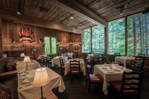 Cozy restaurant interior with wooden walls, tables set for dining, and large windows overlooking a forested area.