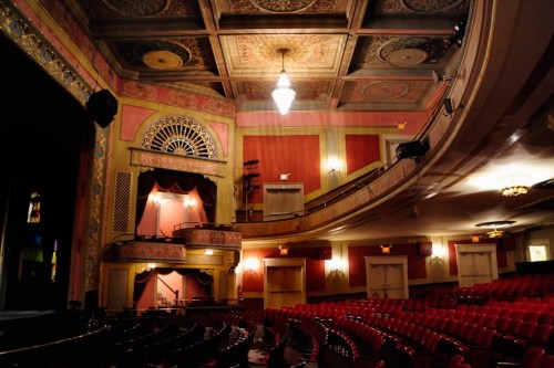 Interior of a vintage theater featuring ornate decor, red seating, and a grand stage with a chandelier overhead.
