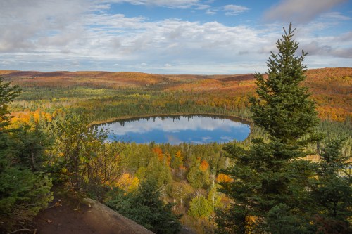A serene landscape featuring a circular lake surrounded by vibrant autumn foliage and evergreen trees under a blue sky.
