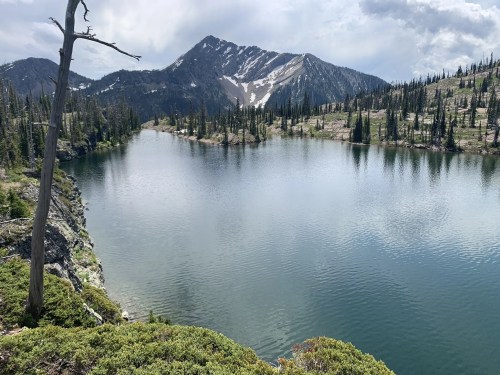 A serene mountain lake surrounded by trees and rocky terrain, with snow-capped peaks in the background under a cloudy sky.