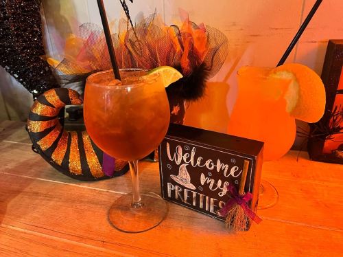 Two colorful cocktails with garnishes sit on a wooden table, alongside a decorative sign that says "Welcome my Pretty."
