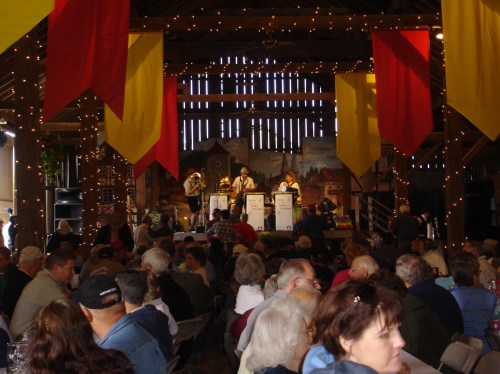 A lively indoor event with a crowd seated at tables, watching musicians perform on stage, decorated with colorful banners.