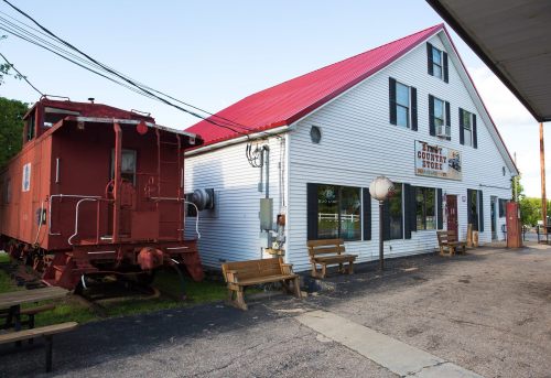 A white building with a red roof and a vintage red train car outside, featuring benches and a sign for a country store.