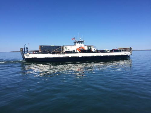A ferry boat glides across calm waters under a clear blue sky, with cargo and equipment visible on deck.